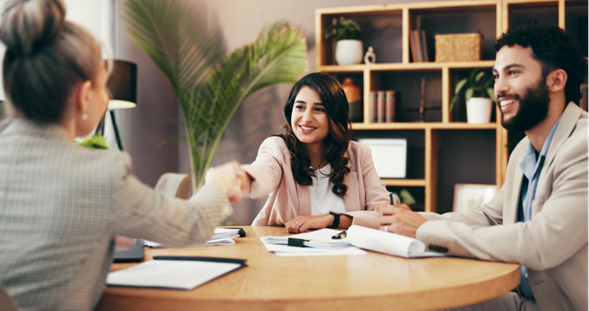 Three professionals in a collaborative meeting around a wooden table, shaking hands and smiling, with notebooks and documents visible, representing business discussion and agreement