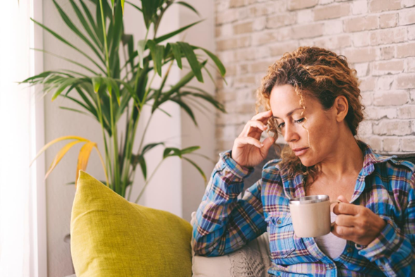 Woman sitting by a window with a green plant, holding a mug, touching her head while appearing thoughtful or fatigued, with brick wall background