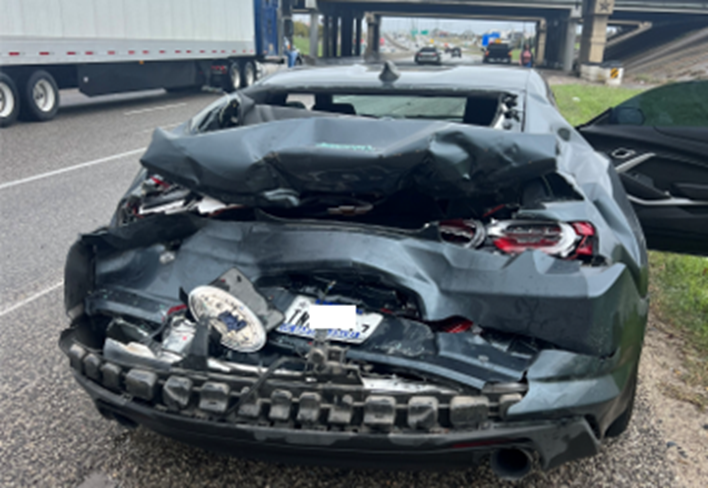 Daños en la parte trasera de un Chevrolet Camaro gris oscuro tras una colisión con un camión en la Interestatal 35, con el maletero abollado, la luz trasera rota y el parachoques desprendido.