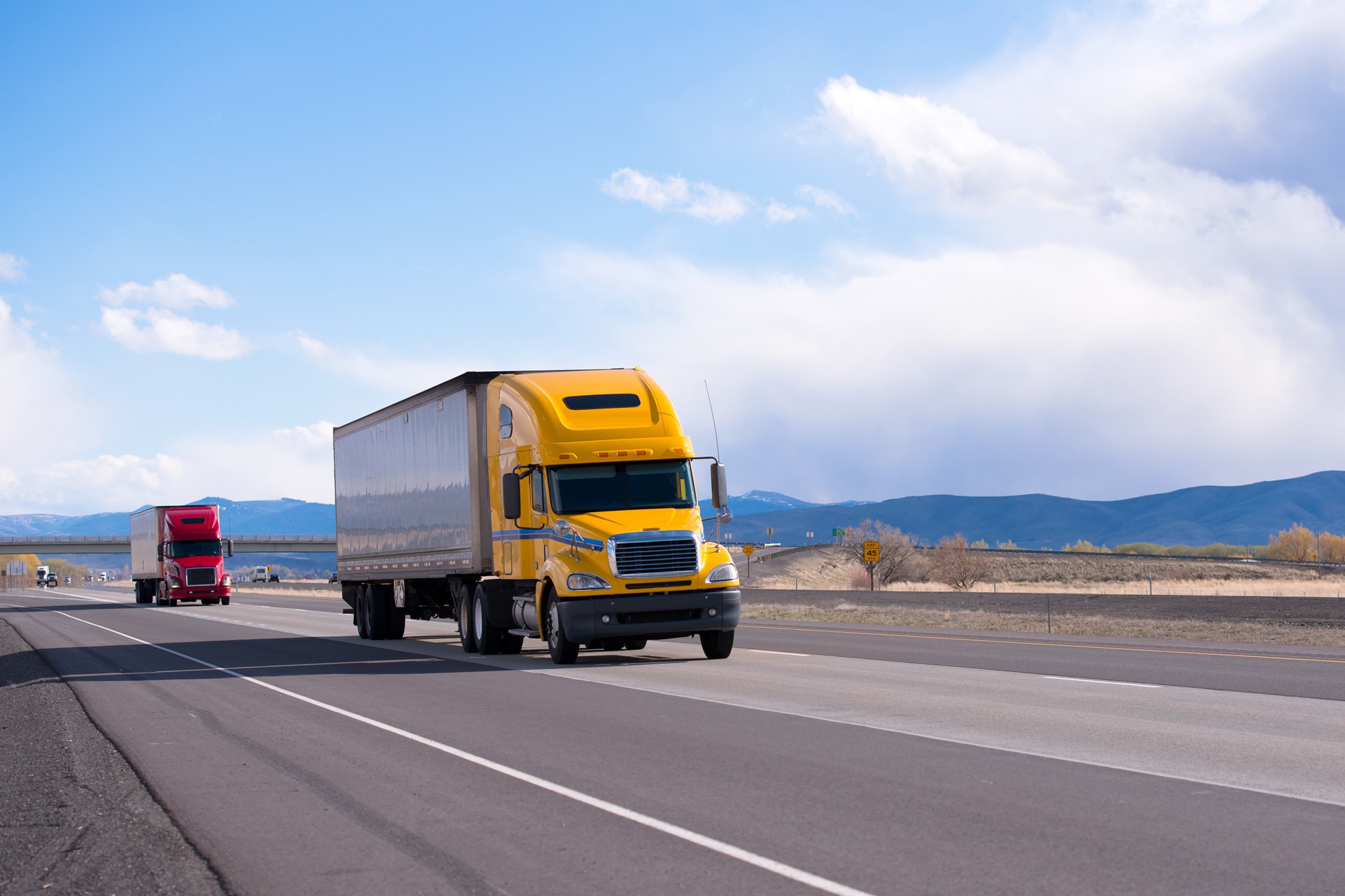 Yellow semi-truck and red commercial truck on Texas highway with mountains in the distance