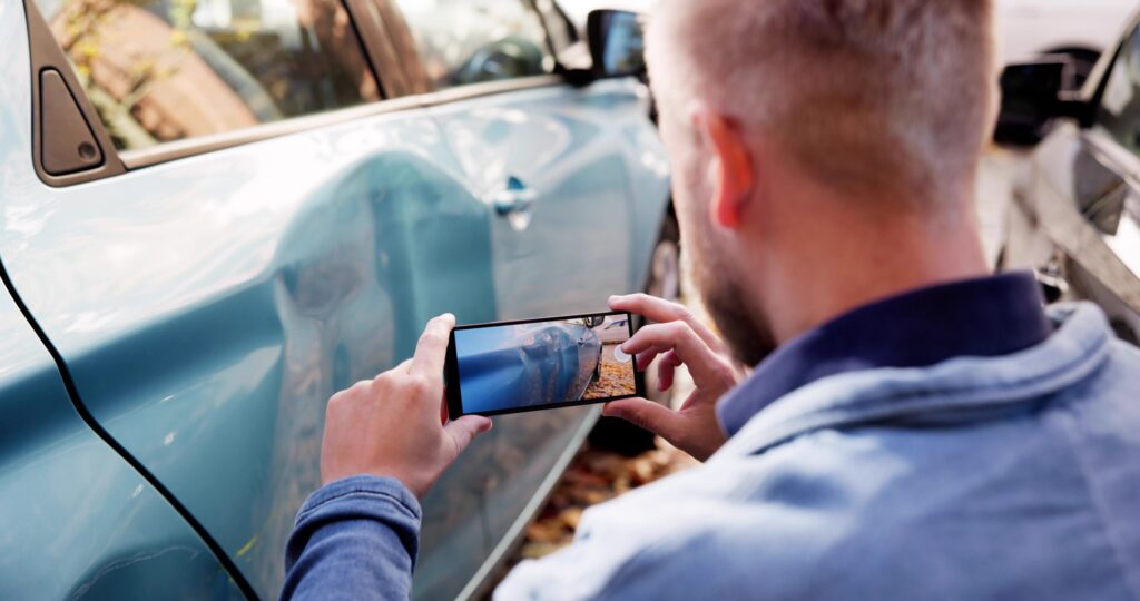 Man using a smartphone to photograph dented door damage on a blue car after a collision