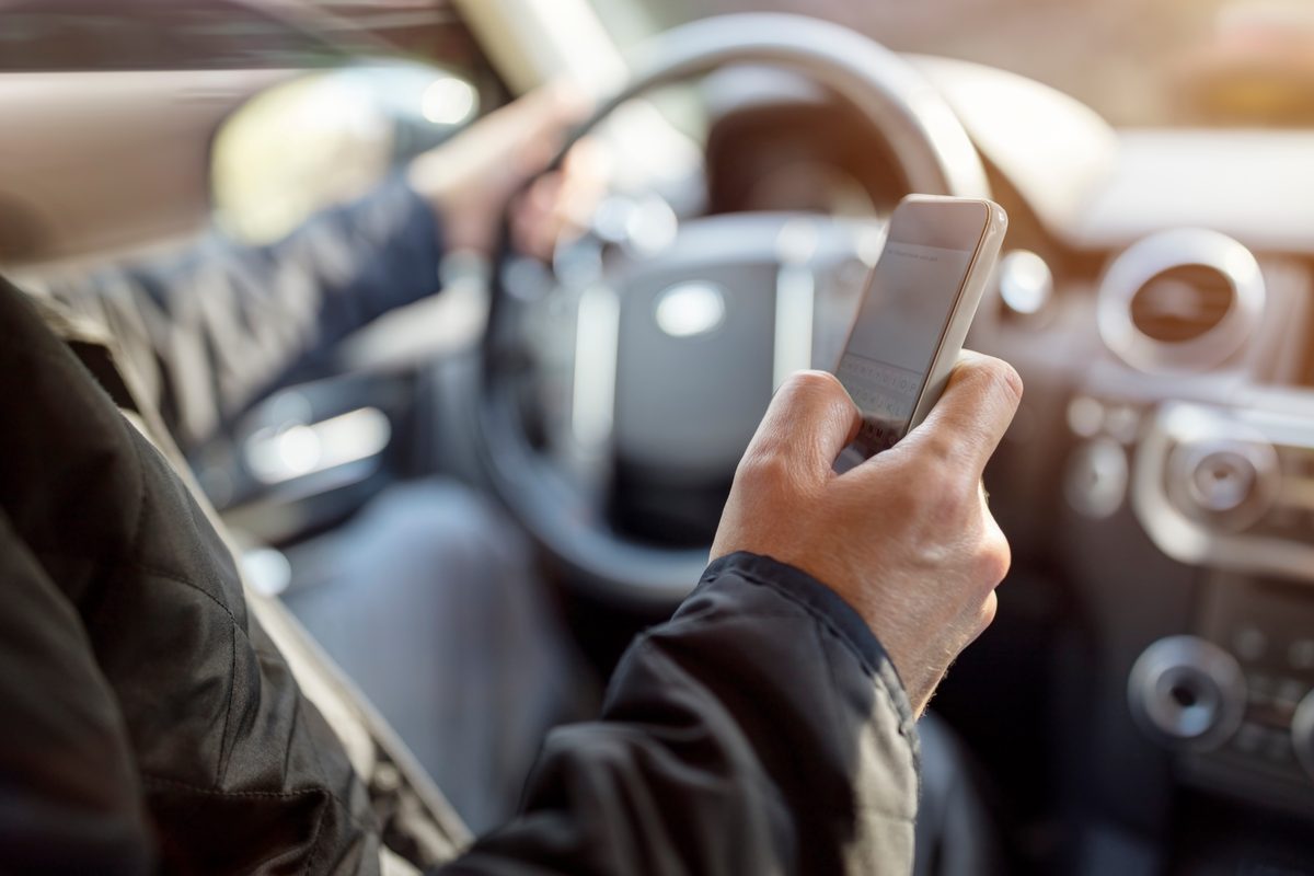 A driver holds a smartphone in one hand while steering with the other, an example of distracted driving